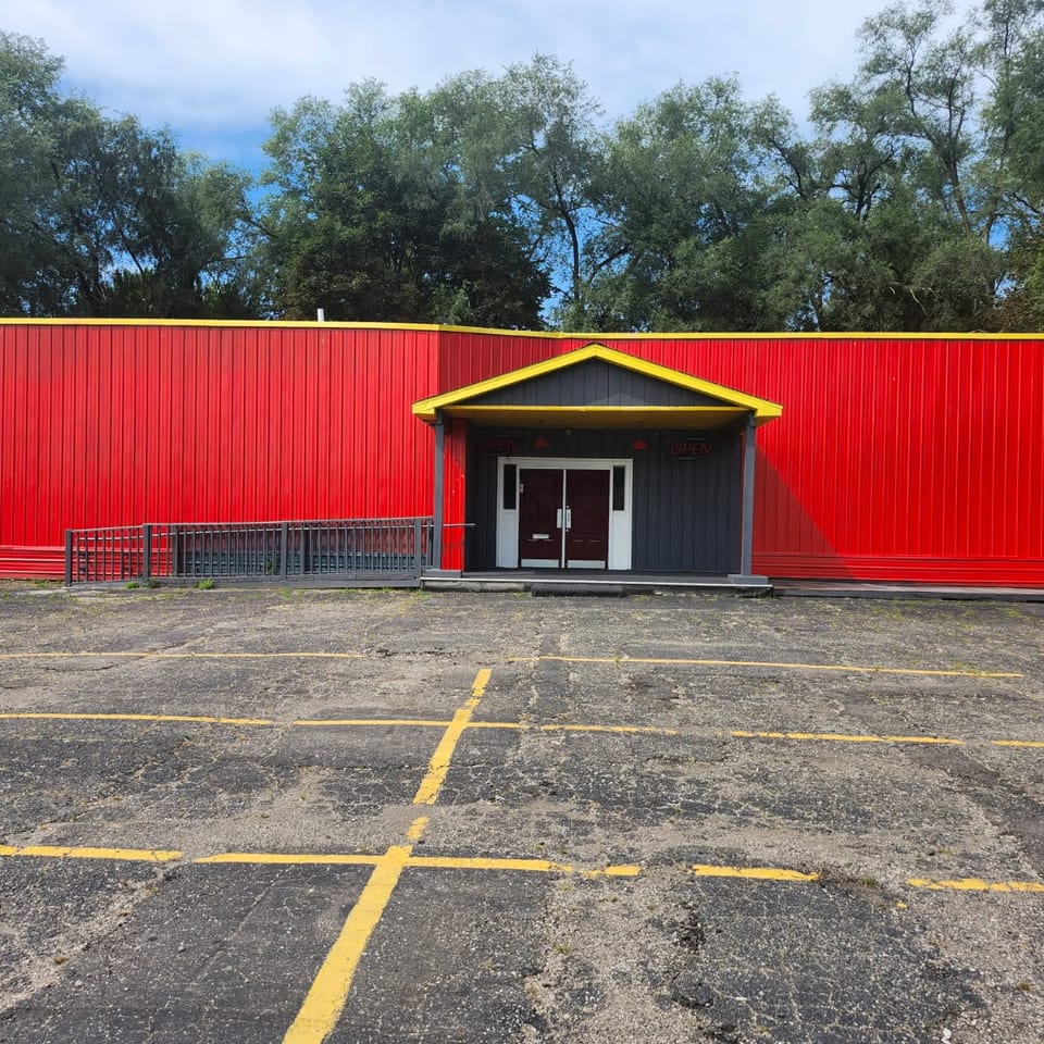 Red restaurant building with yellow trim, covered entrance, accessible ramp, and empty parking lot surrounded by trees.